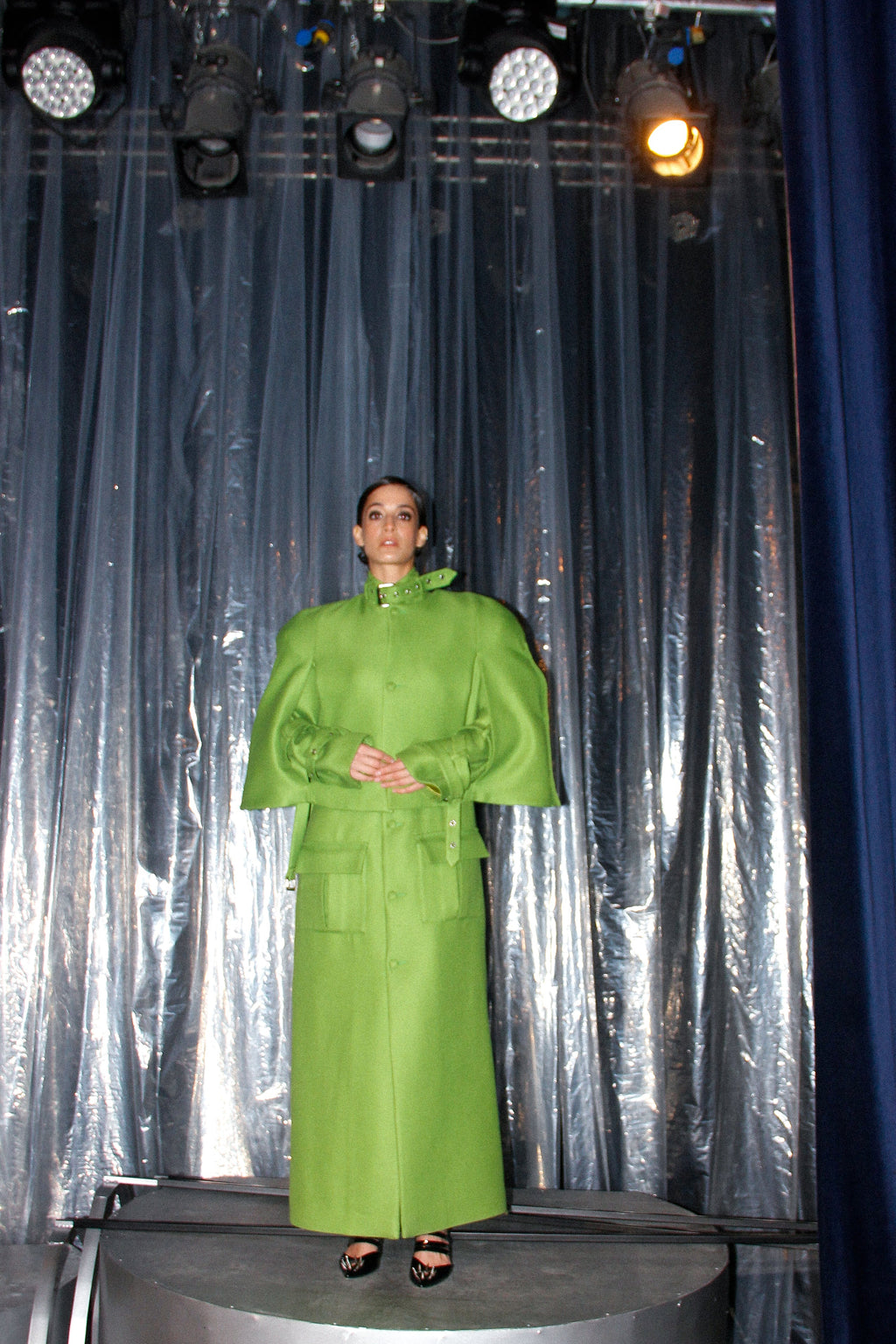 Person wearing a green raincoat standing on a platform with a silver curtain and blue curtain in the background.